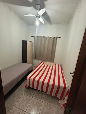 Eine kleine, einfache Unterkunft mit einem Bett, einem Kleiderschrank, einem Deckenventilator und einem Fenster mit Vorhängen.