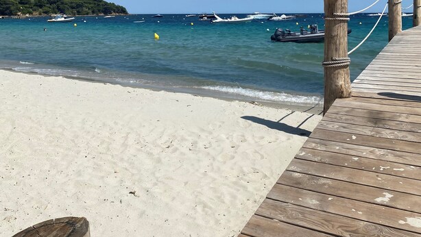 Une jetée en bois mène à une plage de sable fin, où l’eau est d’un bleu limpide et où l’on aperçoit des bateaux au loin.