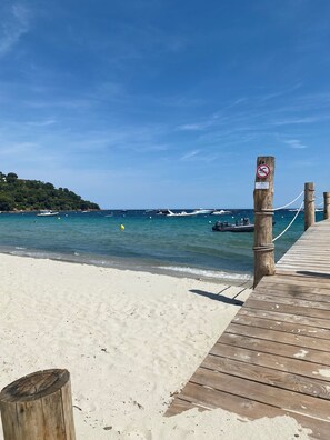 Un muelle de madera que conduce a una playa de arena con agua azul cristalina y barcos a lo lejos.