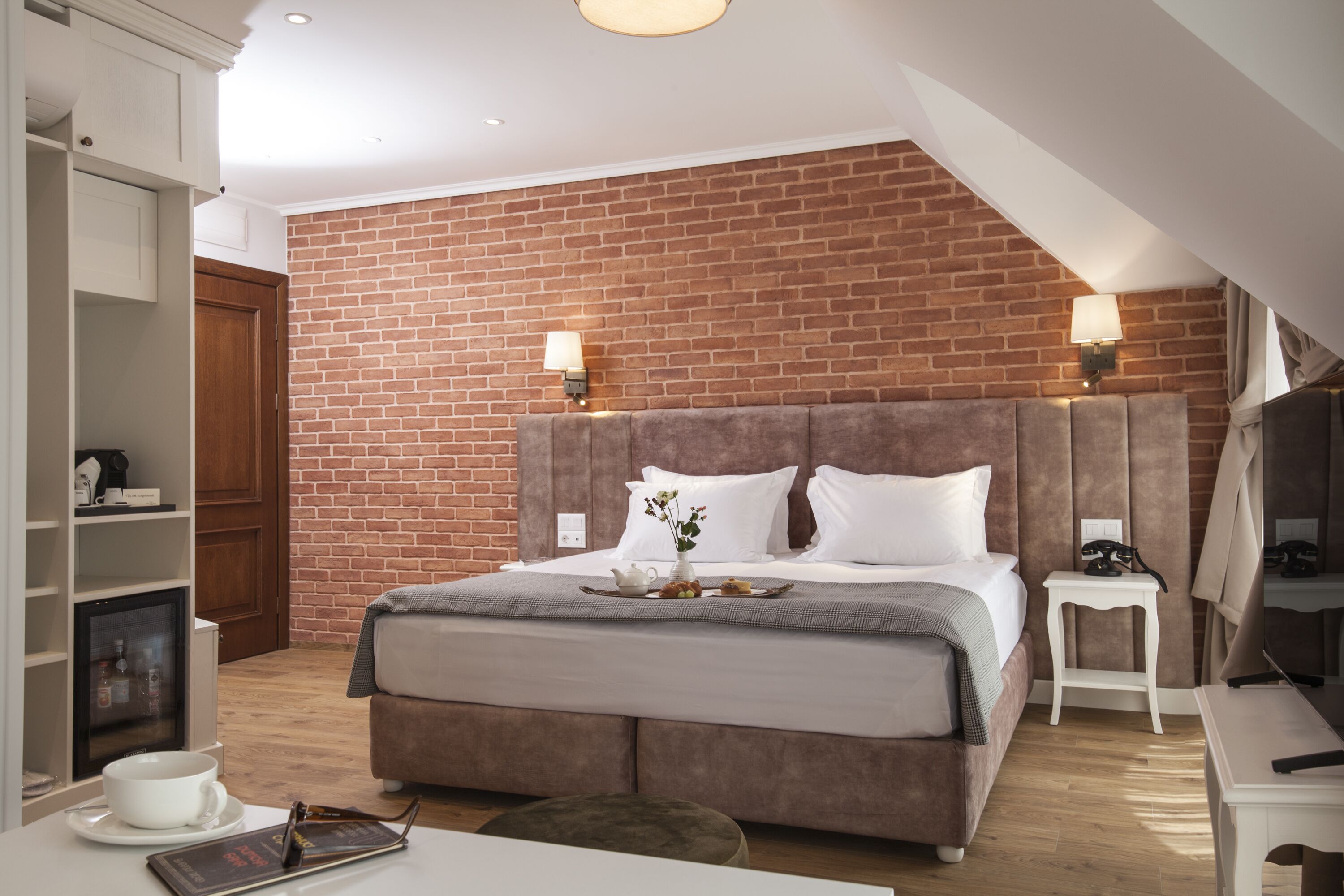 A modern hotel room with a brick wall, a large bed with a headboard, bedside tables, a desk, and a television.