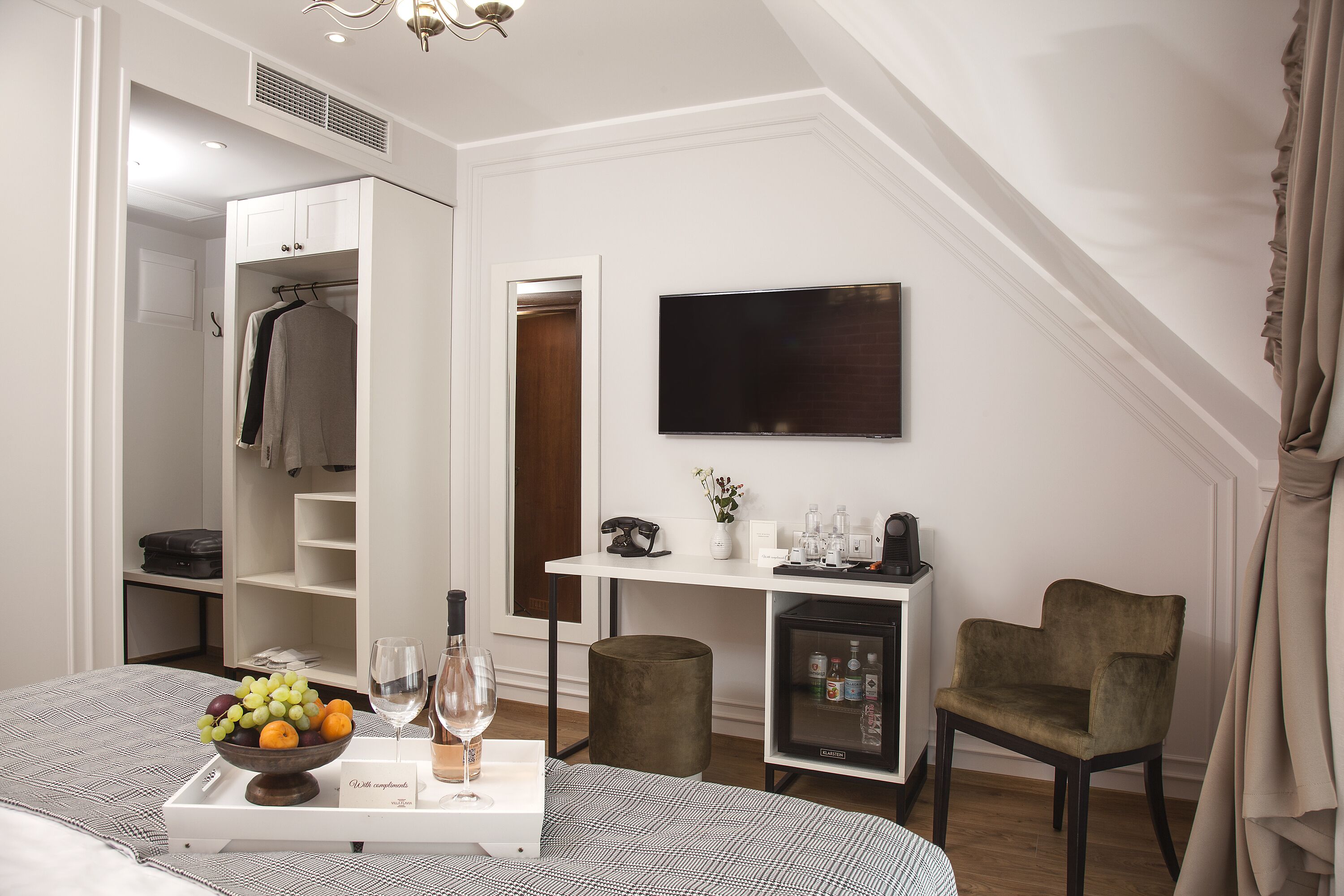 A hotel room with a bed, a bedside table with a fruit bowl and wine, a wardrobe, a desk with a TV, a chair, and a small refrigerator.