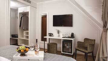 A hotel room with a bed, a bedside table with a fruit bowl and wine, a wardrobe, a desk with a TV, a chair, and a small refrigerator.