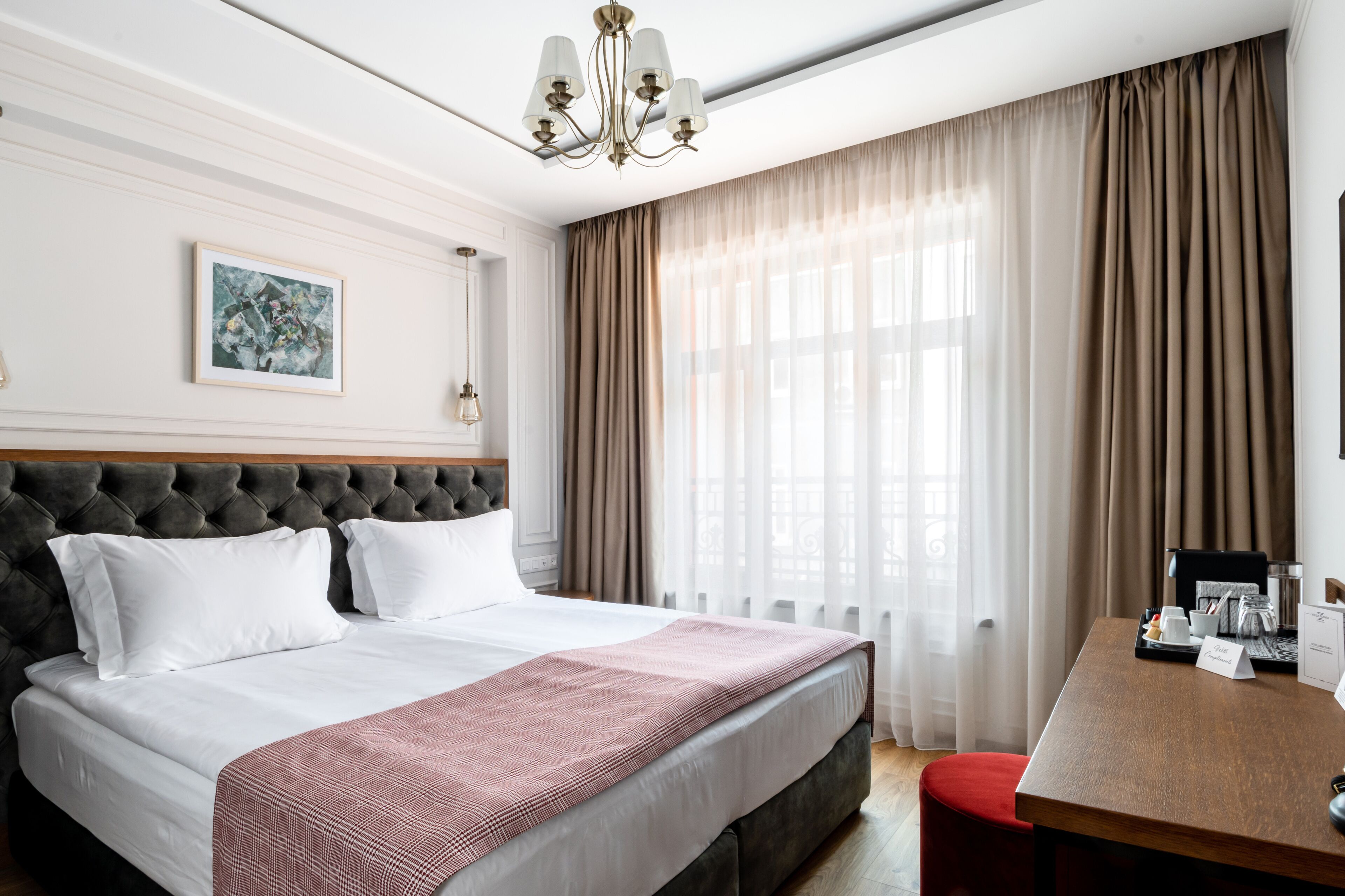 A neatly made bed with white linens and a red blanket, a tufted headboard, a framed picture on the wall, and a desk with a coffee maker and a sign.