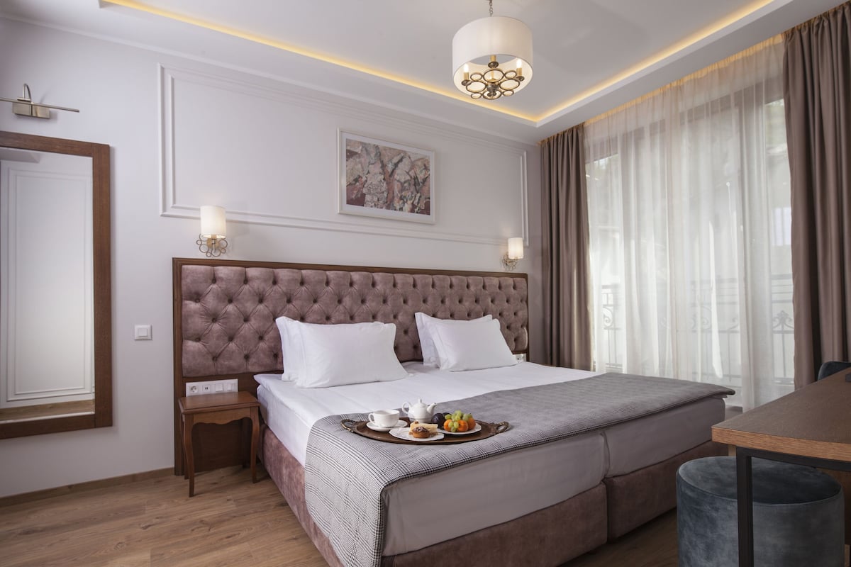 Ein Hotelzimmer mit einem großen Bett, einem Nachttisch mit Lampe, einem Esstisch und einem großen Fenster mit Vorhängen.