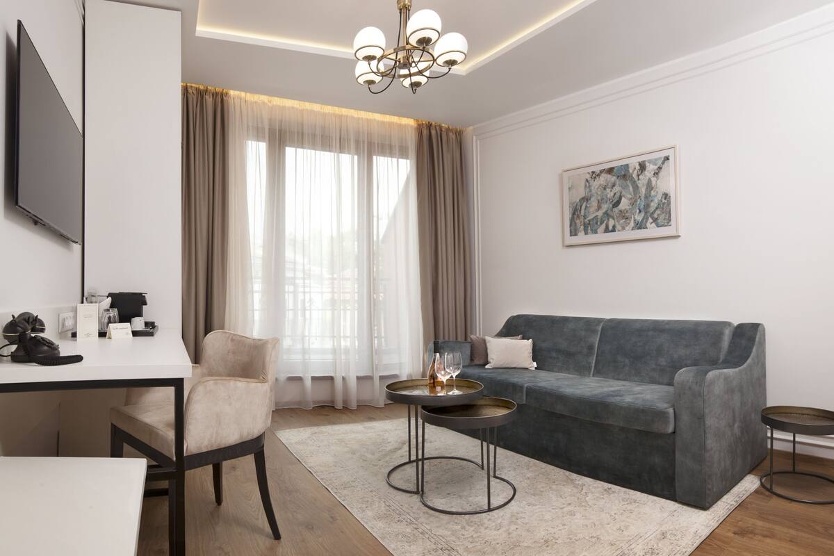 Ein modernes Wohnzimmer mit einem grauen Sofa, einem runden Couchtisch, einem beigen Sessel, einem flachbildfernseher an der Wand und einem großen Fenster mit transparenten Vorhängen.
