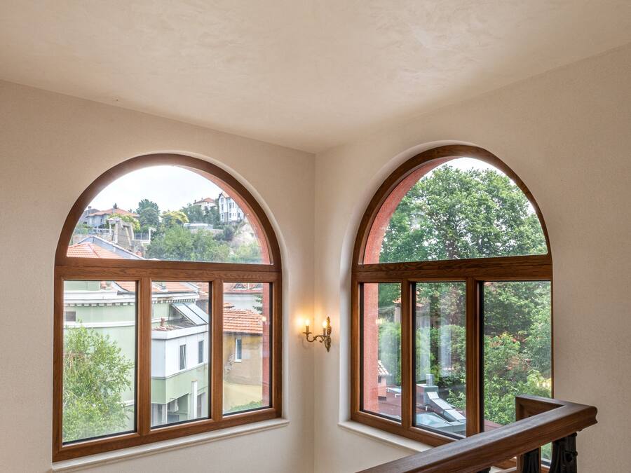 A staircase with ornate black railings and a wooden handrail, leading to a landing with two arched windows overlooking a residential area.
