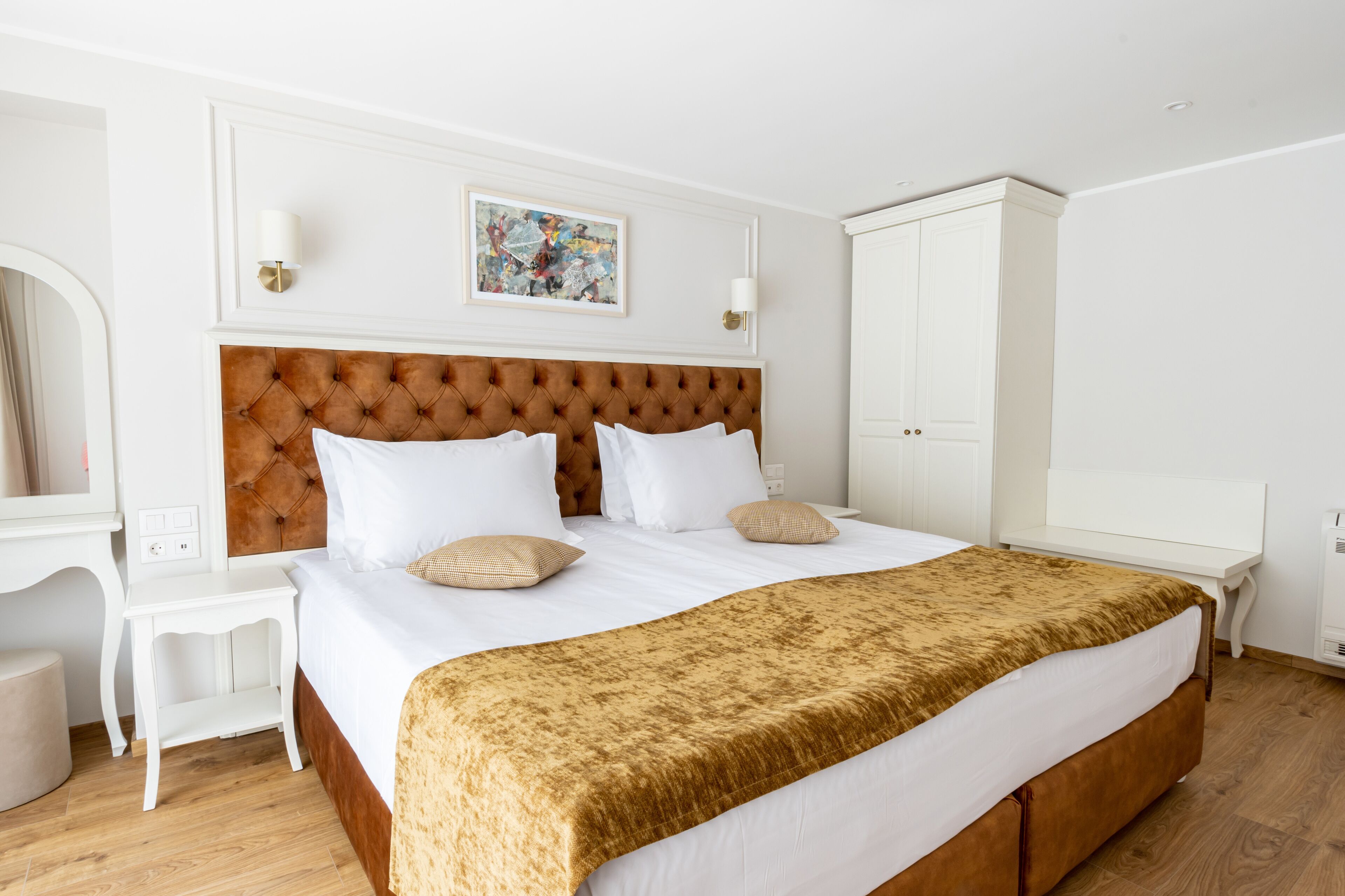 A hotel room with a large bed, white walls, a wooden headboard, a white nightstand, a white dresser, and a framed picture on the wall.