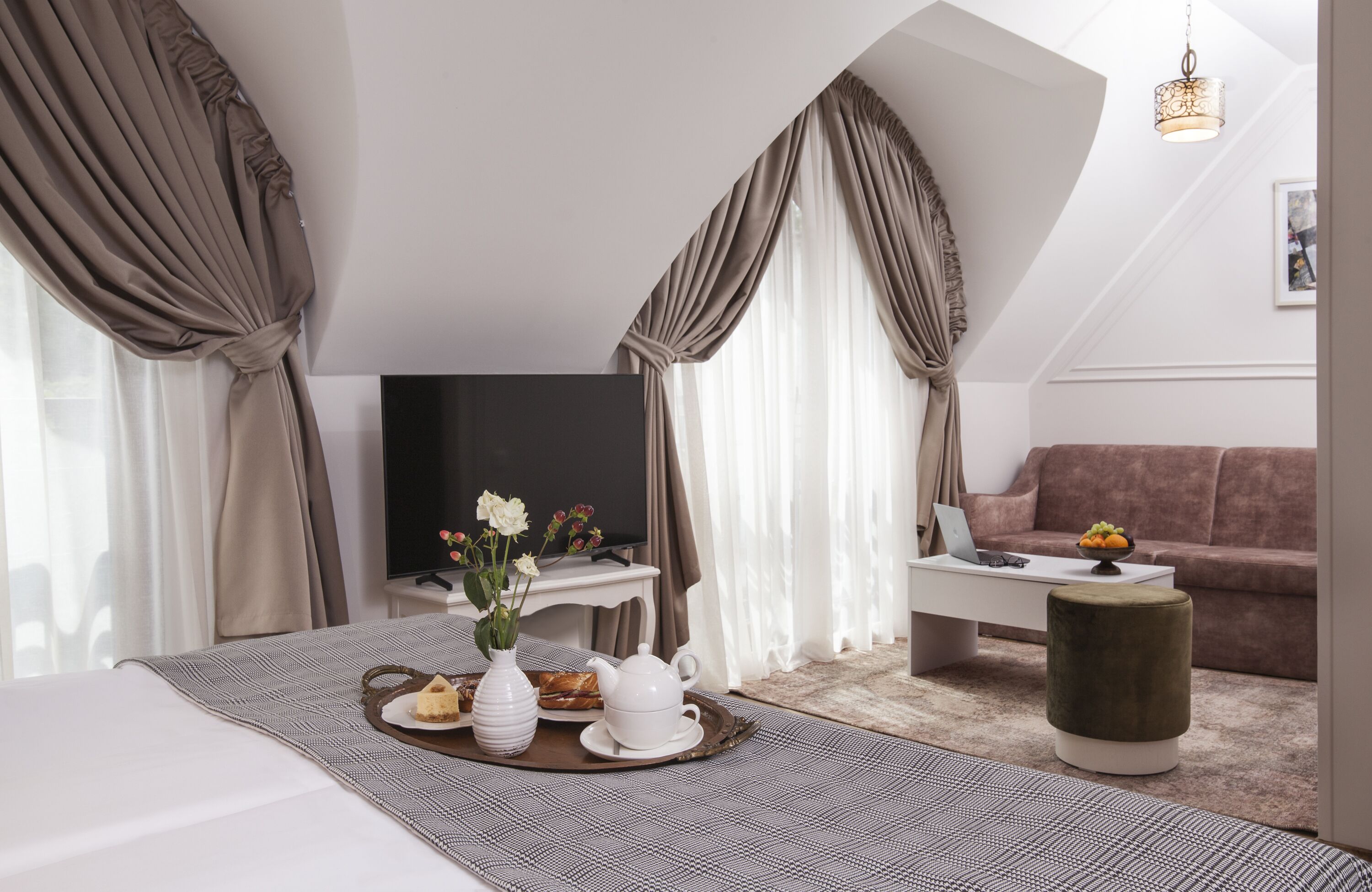 Una habitación de hotel moderna con una cama grande, un televisor de pantalla plana, una mesa de centro, un sofá y una mesa de comedor con una bandeja de pasteles y una tetera.