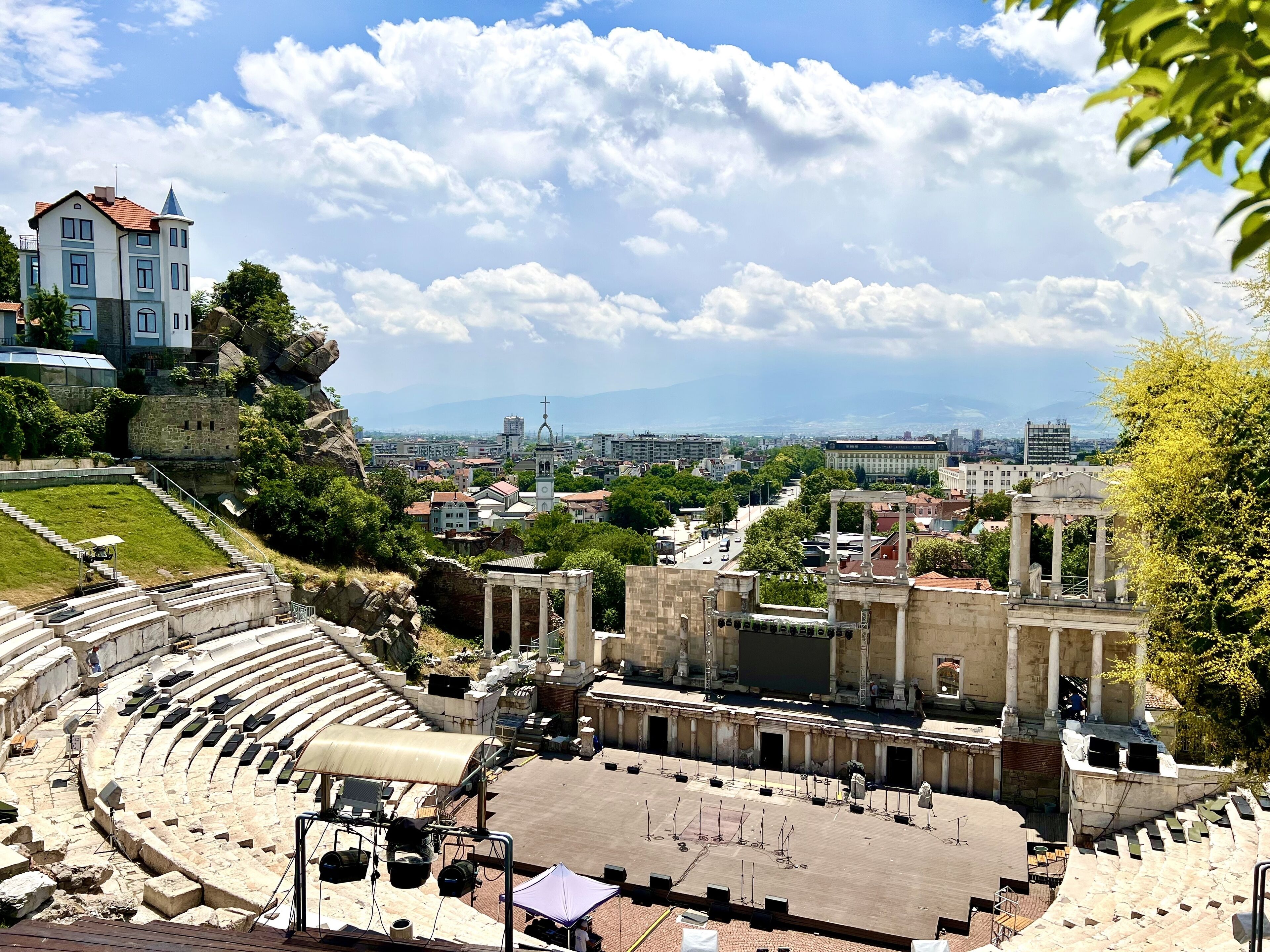 Un anfiteatro antiguo con asientos de piedra y un escenario central, rodeado de edificios históricos y un paisaje urbano al fondo.