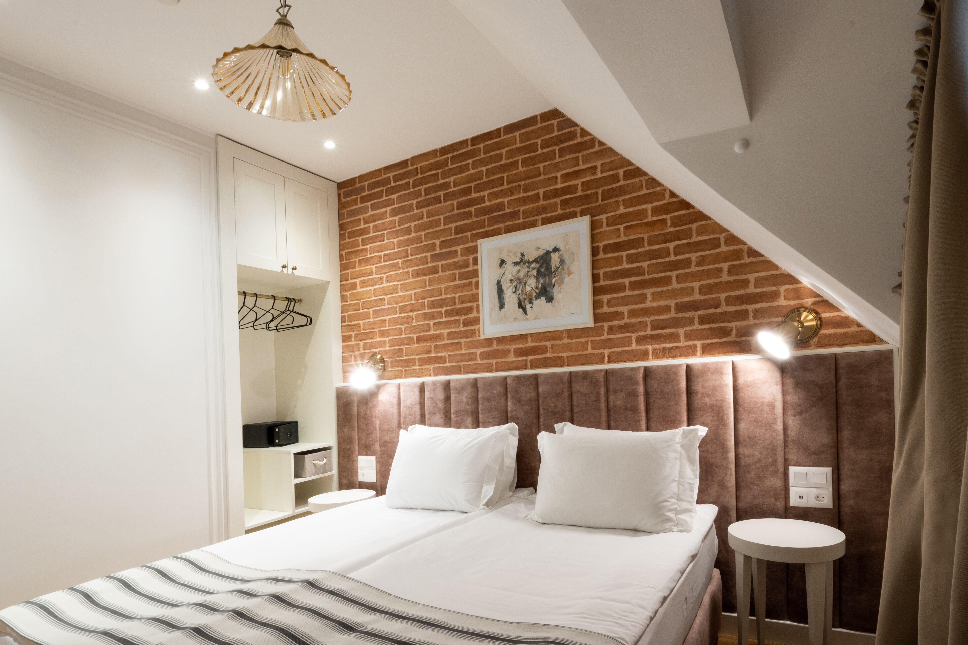 Un dormitorio con pared de ladrillo, cama con ropa blanca, armario y mesita de noche con lámpara.