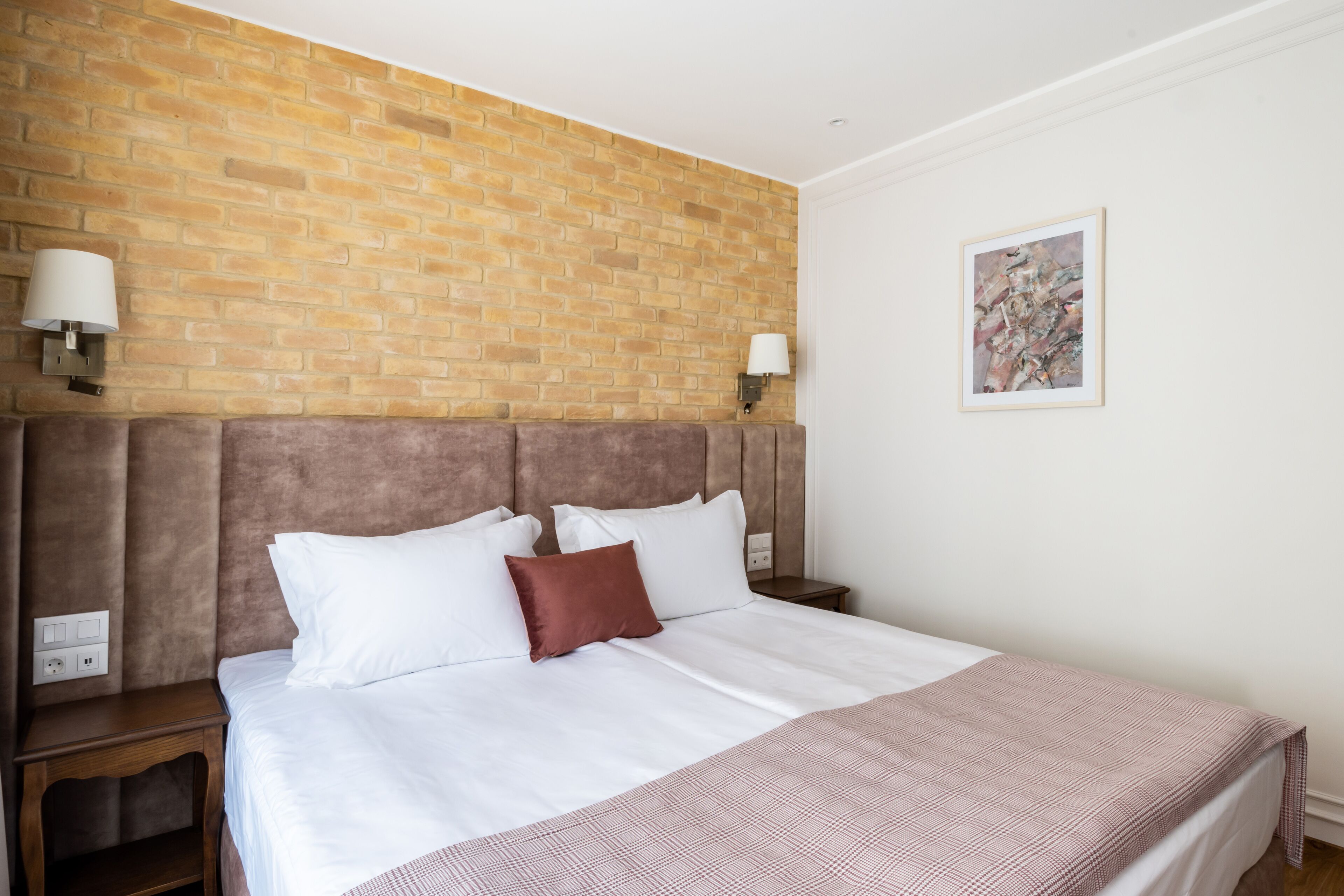 Una habitación de hotel con pared de ladrillo, una cama grande con ropa blanca, cabecera marrón, dos lámparas de noche y un cuadro abstracto enmarcado en la pared.