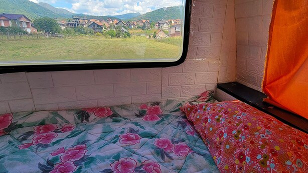 A train compartment with a bed, a window with a view of houses and mountains, and a brick wall.