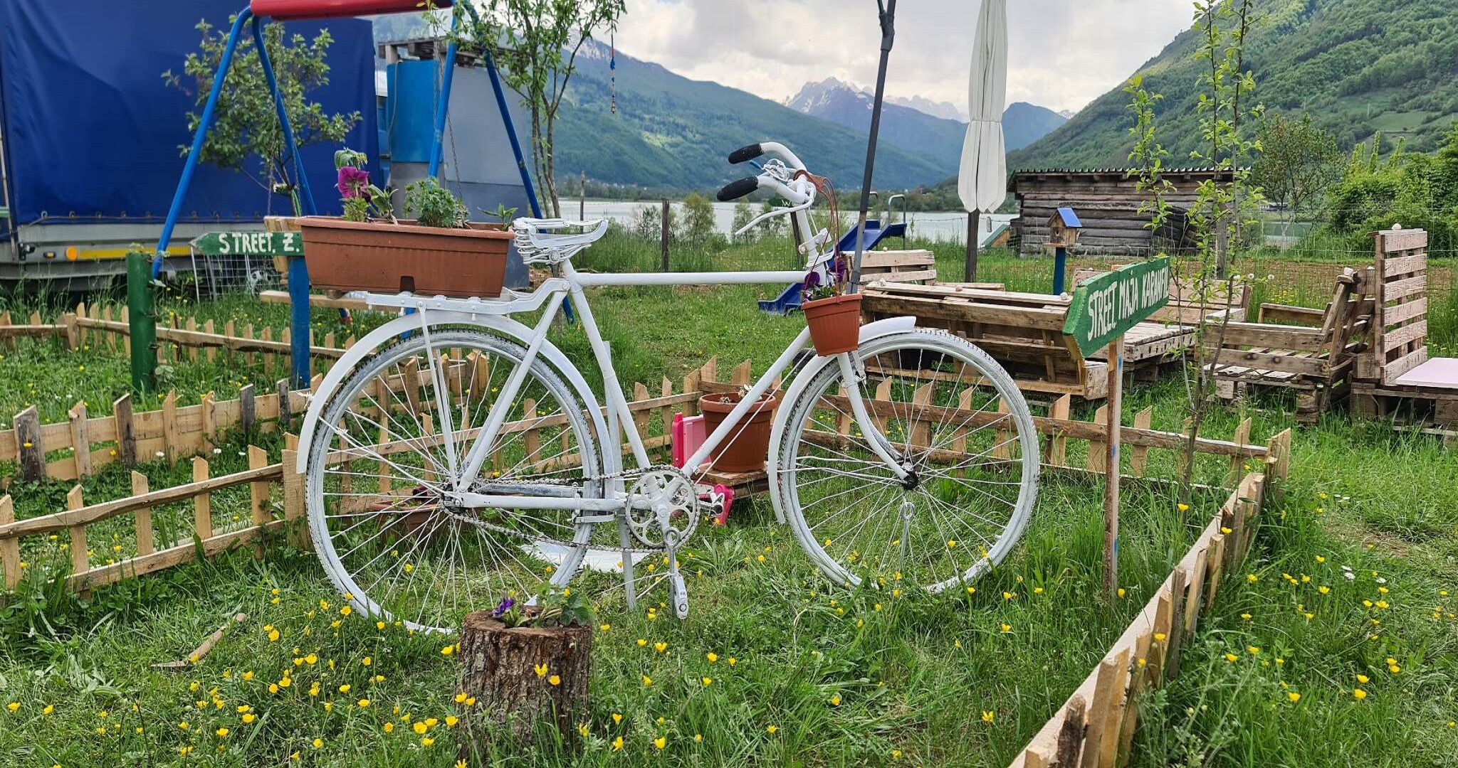 Uma bicicleta branca estacionada em área gramada, com cerca de madeira, estrutura de madeira e uma placa com a inscrição 'STREET'.