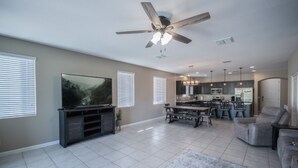A spacious living room with a flat-screen TV, a ceiling fan, and a dining area with a kitchen in the background. - Like New Home Private pool ,New furnishings, 20 mins to State Farm stadium  (Buckeye)