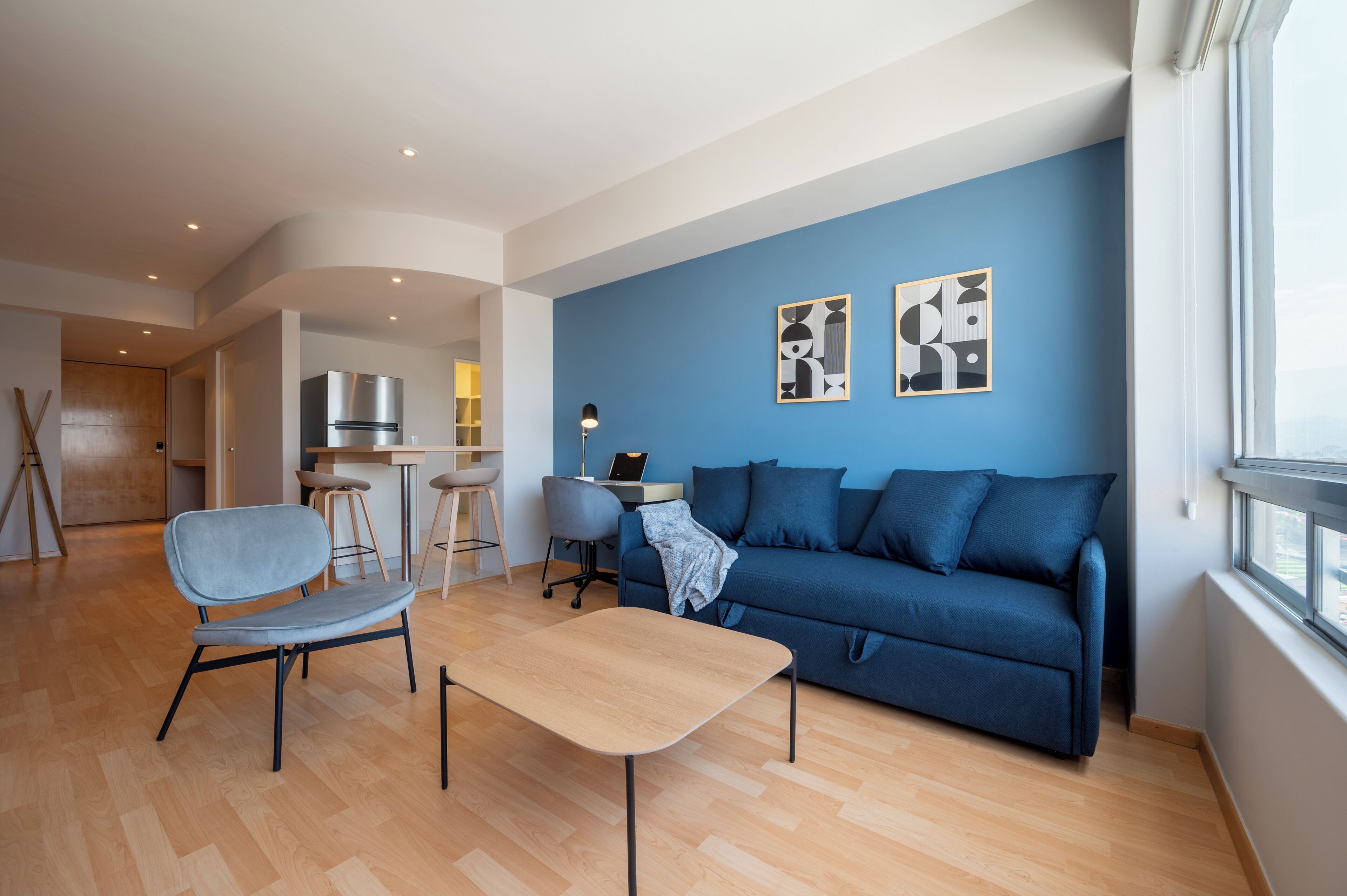 Un salon moderne avec un canapé bleu, une table basse en bois et un fauteuil gris. On y trouve également un coin cuisine avec un réfrigérateur en acier inoxydable et un ordinateur portable posé sur le comptoir.