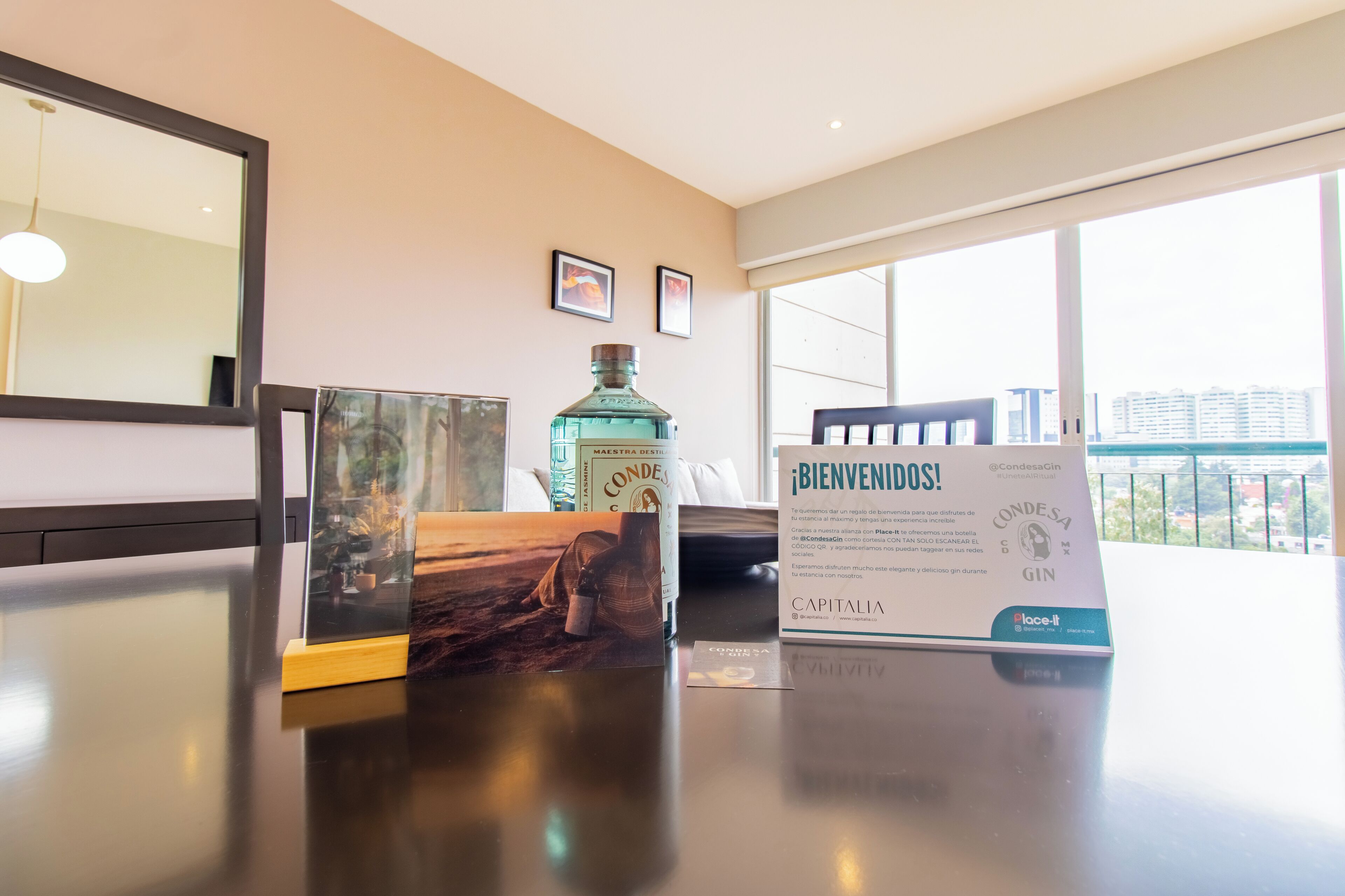 A hotel reception area with a large window, a framed welcome sign, a bottle of gin, and a framed picture on the counter.
