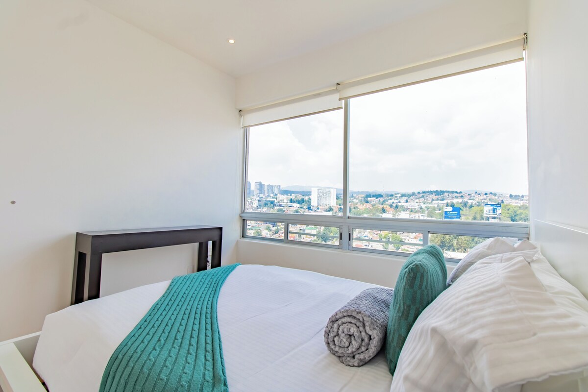 Ein Hotelzimmer mit großem Fenster, einem Bett mit weißer Bettwäsche, einer türkisfarbenen Tagesdecke und einer aufgerollten grauen Decke.