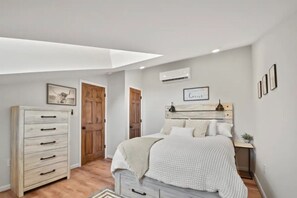 A bedroom with a bed, a dresser, a wooden door, and a wall-mounted air conditioner.