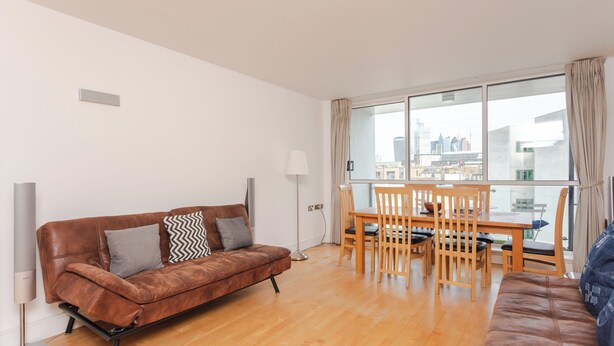 A modern living room with a brown leather sofa, a dining table with chairs, and a large window offering a city view. - Cosy and Spacious 3 BD flat on Bermondsey St (London)