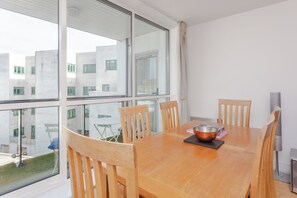 Un coin repas avec une table et des chaises en bois, un saladier, et une vue sur un bâtiment à travers de grandes fenêtres.