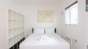Une chambre à coucher avec un lit, du linge de lit blanc, une œuvre d’art représentant une carte du monde, une fenêtre avec des stores et une armoire blanche dotée de plusieurs étagères.