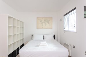 Une chambre à coucher avec un lit, du linge de lit blanc, une œuvre d’art représentant une carte du monde, une fenêtre avec des stores et une armoire blanche dotée de plusieurs étagères.