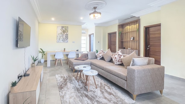Un salon moderne avec un canapé, une table basse et un téléviseur. On y trouve également un coin repas avec des chaises et une table. La pièce est équipée d’un ventilateur de plafond et possède une porte en bois.