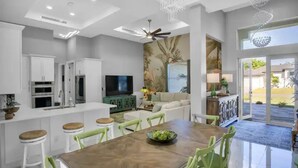 A modern kitchen with white cabinets, a dining area with a wooden table and green chairs, and a large window with a view of the outdoors. - Luxurious SW Florida Pool Home (Cape Coral)