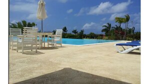 Un área de piscina con sillones de descanso y una mesa, rodeada de palmeras y un cielo despejado.