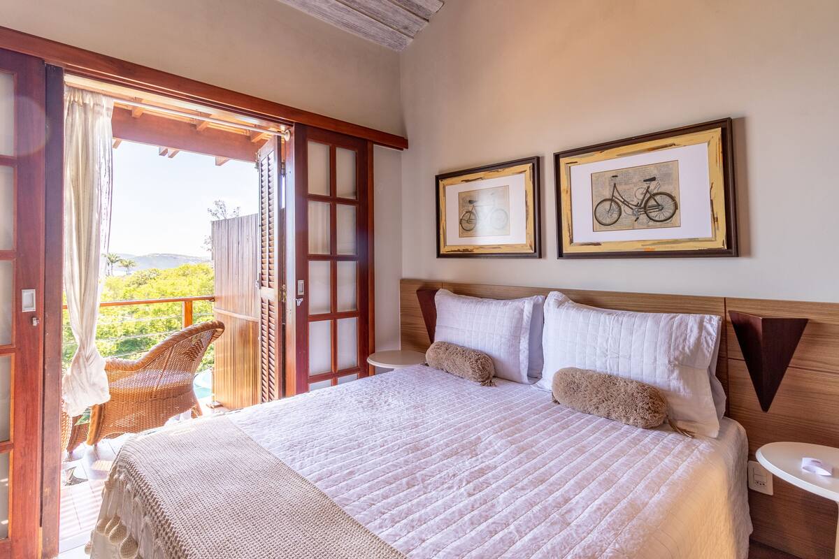 A bedroom with a large bed, a wooden headboard, two framed pictures, a small bedside table, and a wicker chair by a sliding glass door.
