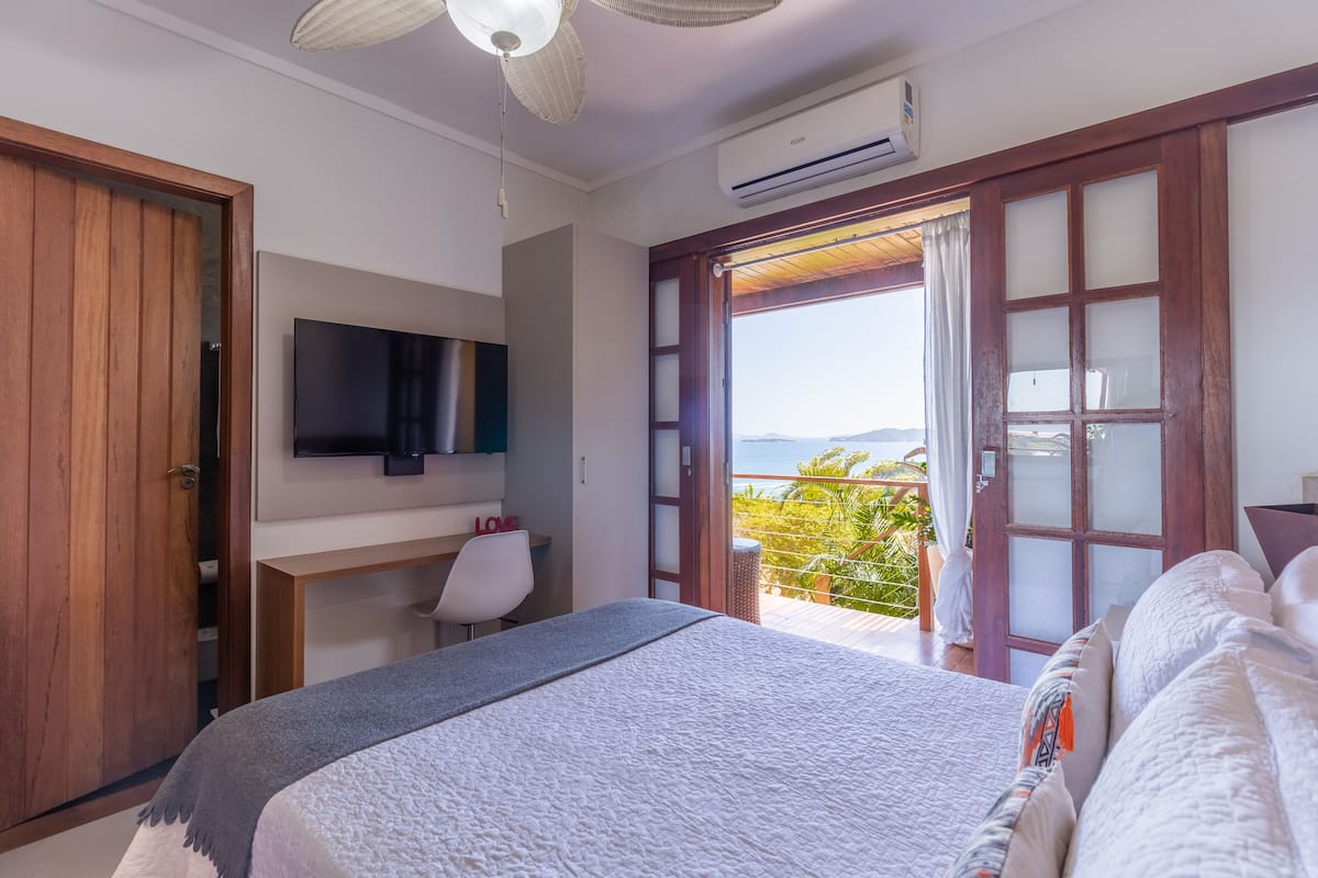A bedroom with a bed, a desk, a chair, a TV, and a balcony with a view.