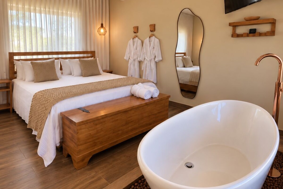 A modern hotel room with a large bed, a sitting area with a wooden bench, a freestanding bathtub, and a mounted TV.