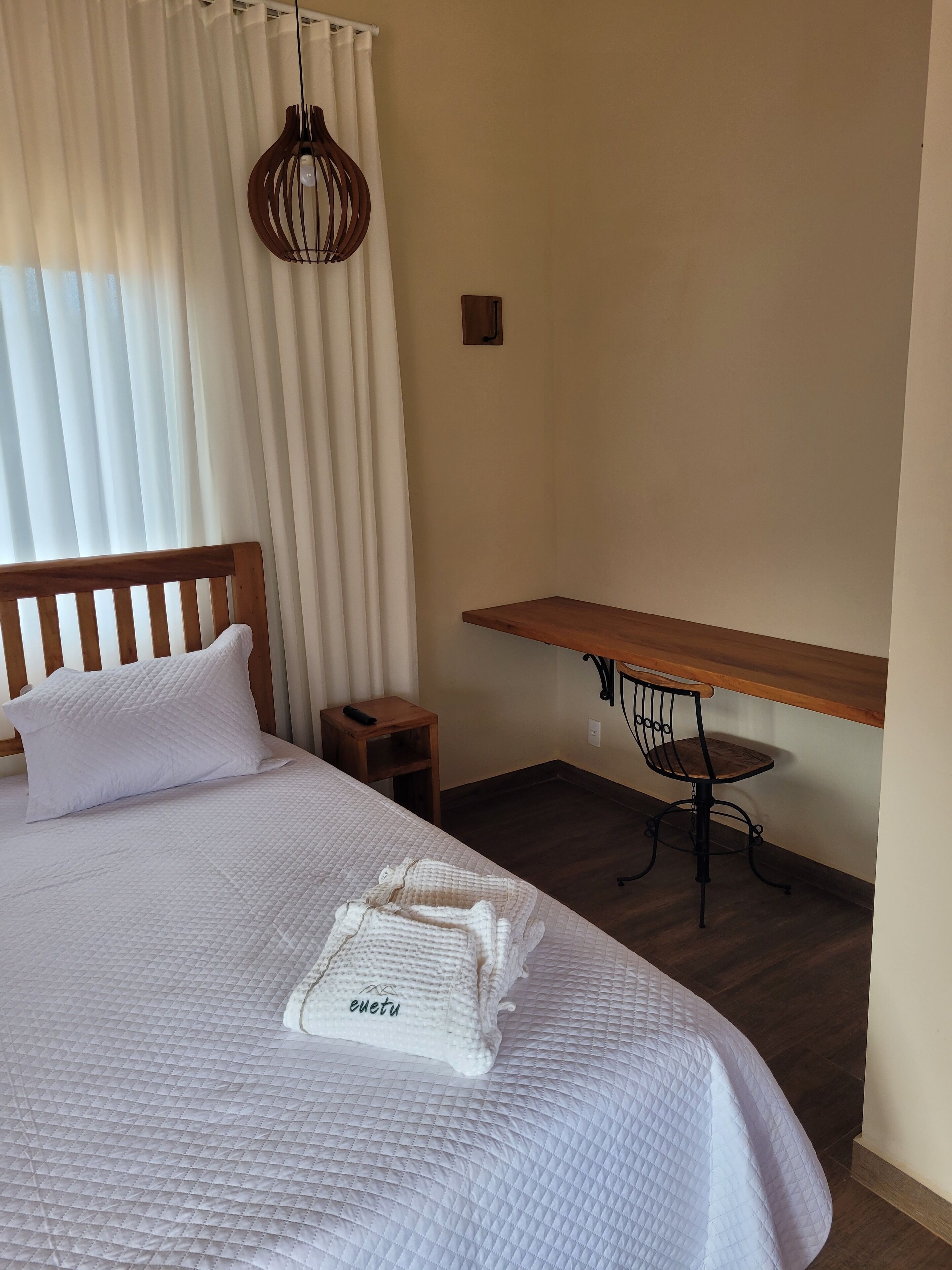 A hotel room with a bed, a wooden nightstand, a desk with a chair, and a hanging light fixture.