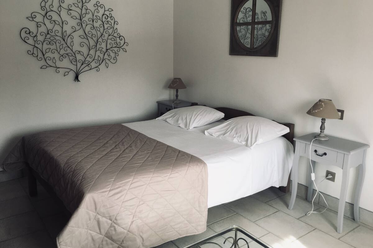 Un lit bien fait, recouvert d’une courtepointe, une table de chevet avec une lampe, un élément de décoration murale et une fenêtre agrémentée d’un cadre décoratif.