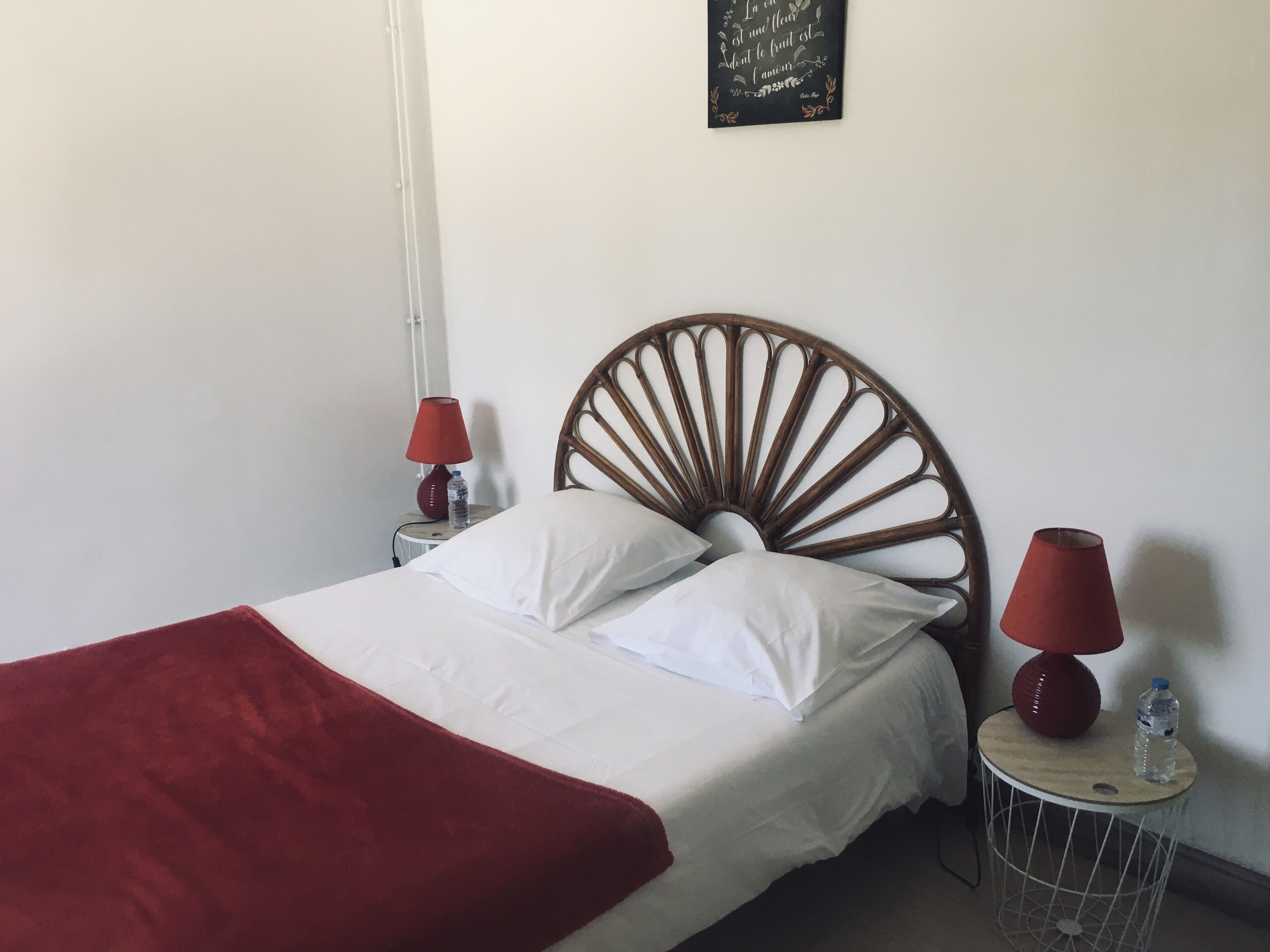 Una cama con cabecera de madera, ropa de cama blanca y una manta roja. Hay dos mesitas de noche con lámparas y botellas de agua.