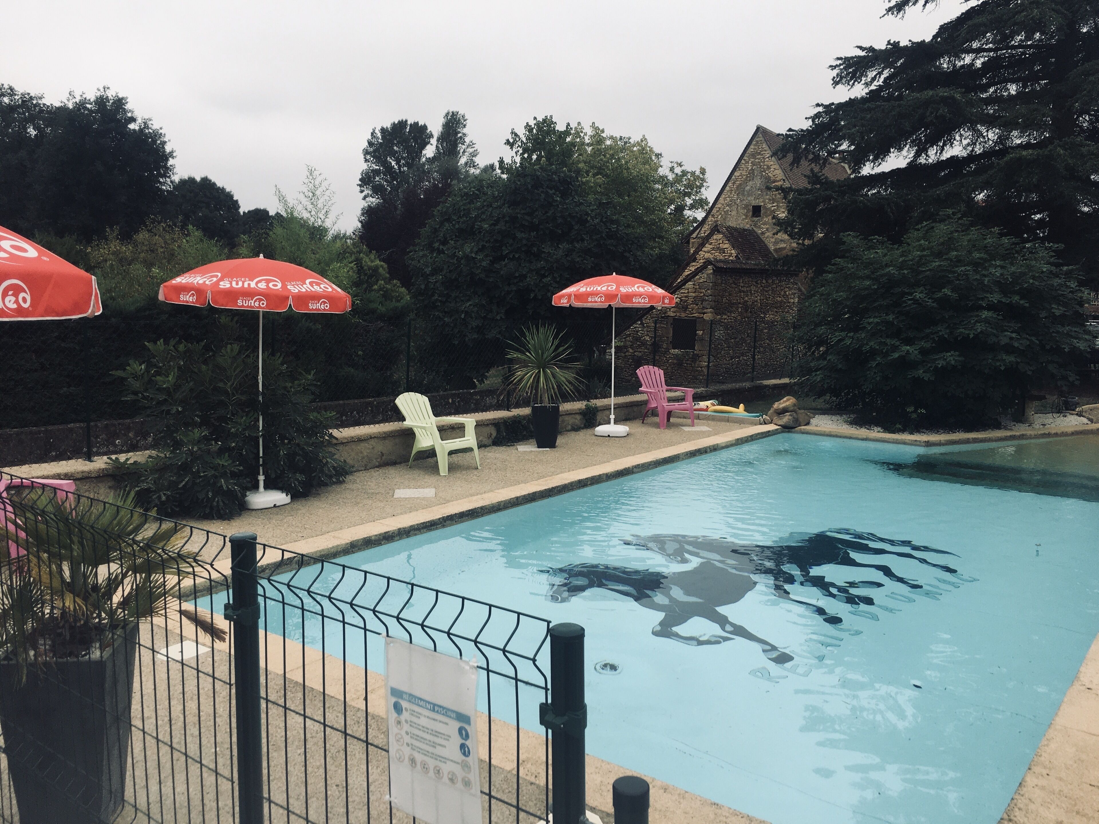 Une piscine entourée d’une clôture, deux parasols rouges et, au fond, un bâtiment en pierre.