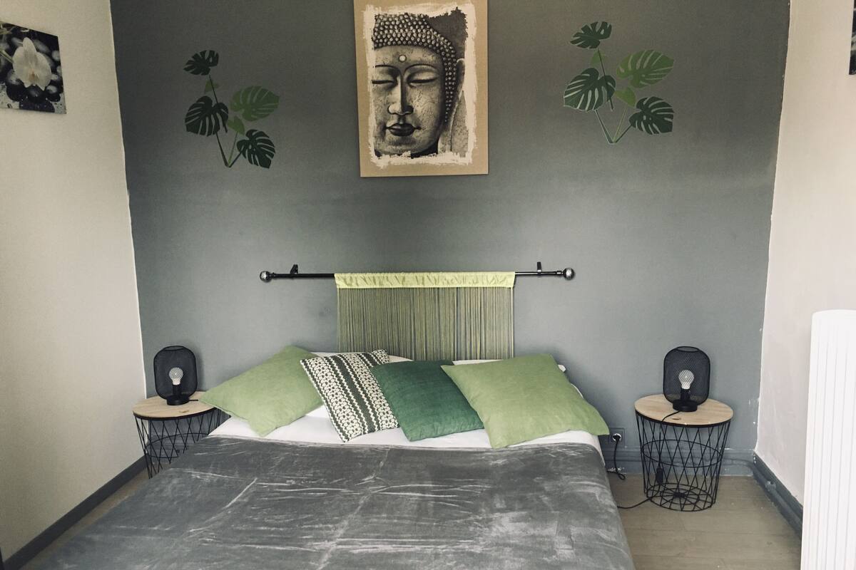 Une chambre à coucher comprenant un lit, deux tables de chevet, un haut-parleur fixé au mur, un rideau et une sculpture représentant une tête de Bouddha.