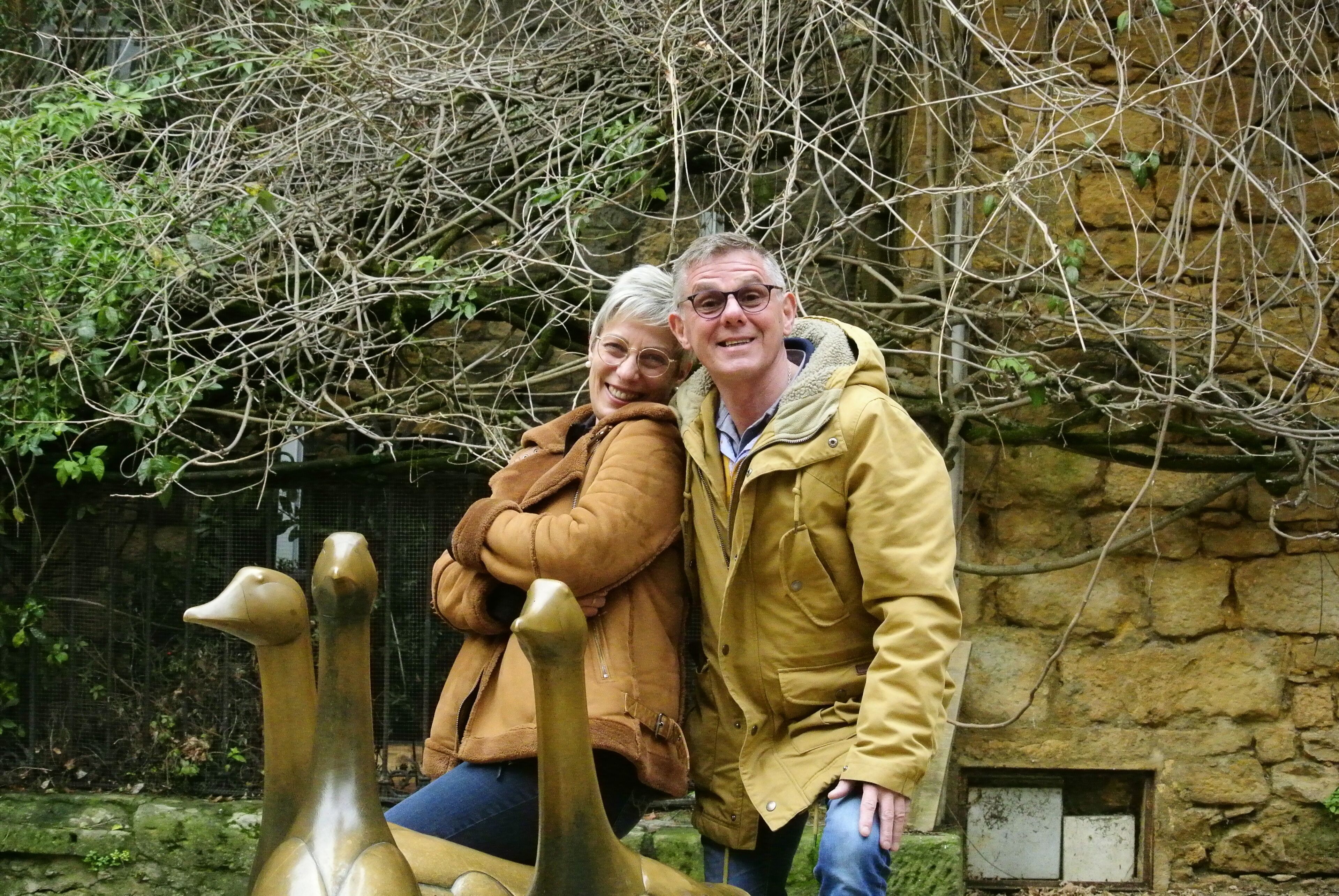 Un couple se tient à côté d’une sculpture en bronze représentant deux autruches, dans un décor extérieur avec un mur de pierre et une végétation luxuriante en arrière-plan.