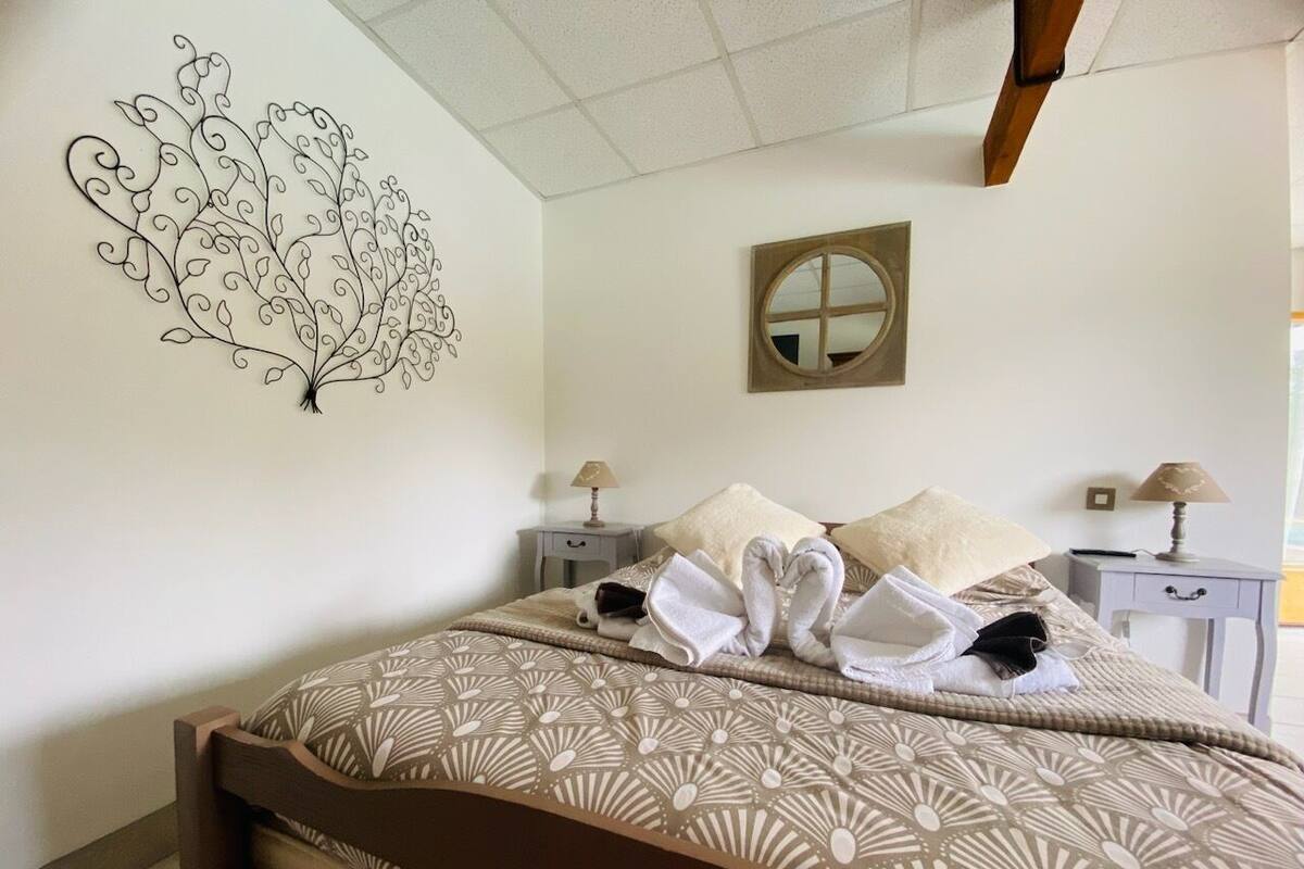 Un lit bien fait, avec des serviettes disposées en forme de cœur, une œuvre d’art murale et un miroir rond placé au-dessus d’une table de chevet.