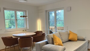 A modern living room with a grey sofa, brown chairs, a round wooden table, and large windows offering a view of greenery. - Cabinet Riedersteinblick in Rottach-Egern am Tegernsee  (Rottach-Egern)