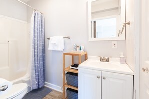 A bathroom with a bathtub, a toilet, a sink with a mirror, and a shelf with toiletries. - New Hot Tub, Surf Studio, Easy Walk to Beach! (Carolina Beach)