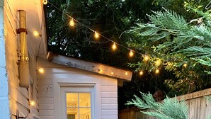 Un petit abri en bois blanc, doté d’une porte et de fenêtres, entouré d’escaliers en pierre et d’une clôture en bois. À l’extérieur, on trouve deux chaises en osier et une table, le tout illuminé par des guirlandes lumineuses.