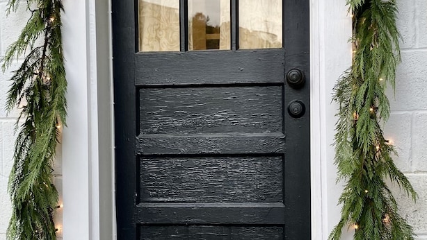Une porte noire ornée d’une fenêtre, flanquée de guirlandes de verdure et de guirlandes lumineuses, qui mène à un bâtiment blanc doté de fondations en pierre.