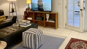 Un salon comprenant un canapé, un pouf, une table basse, un téléviseur, une bibliothèque et une porte avec une fenêtre.