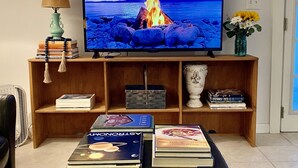 Un salon avec un téléviseur diffusant des images d’un incendie, un canapé, une table basse avec des livres et une lampe.