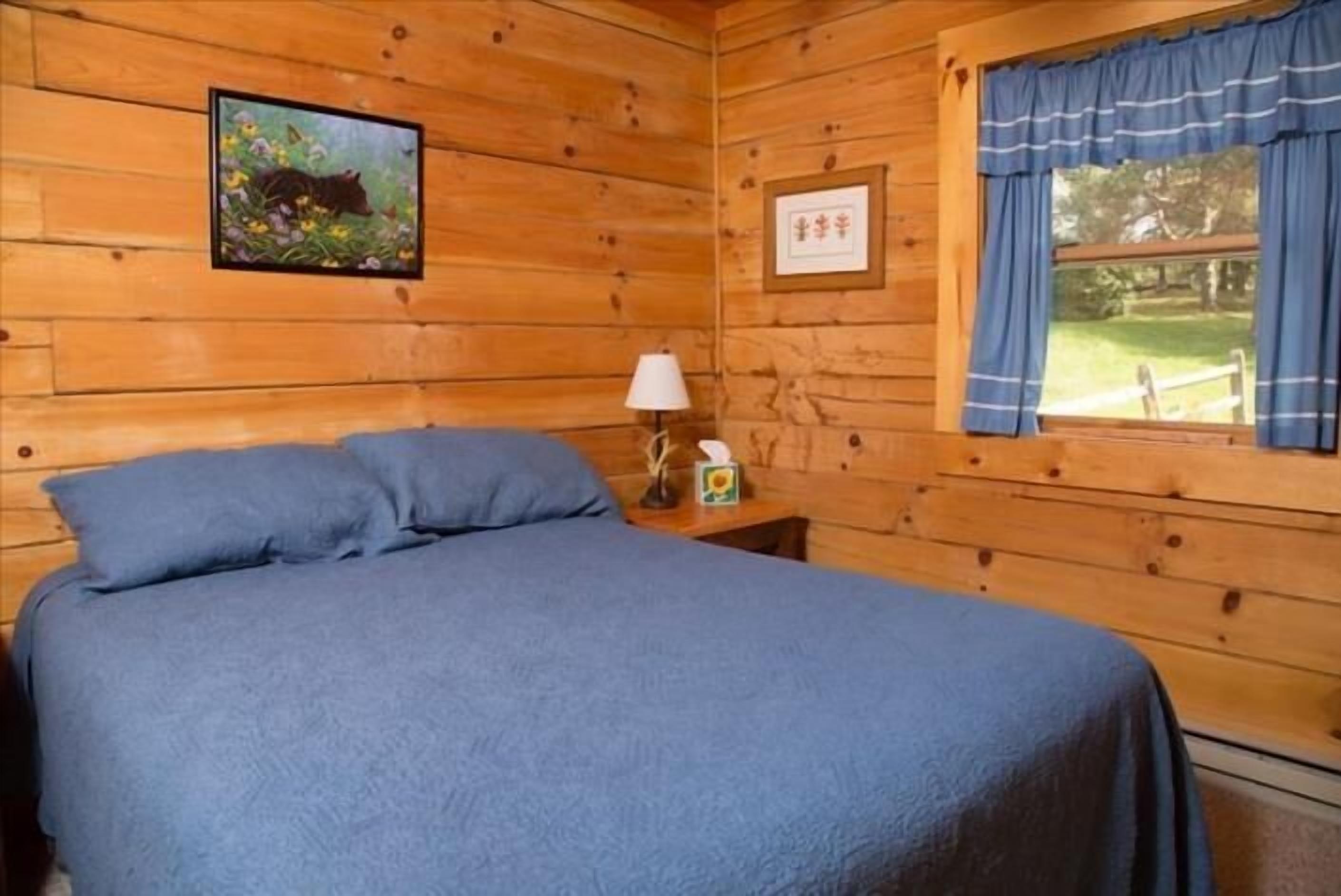 A wooden cabin room with a bed, a framed picture of a bear, a lamp, and a window with curtains.