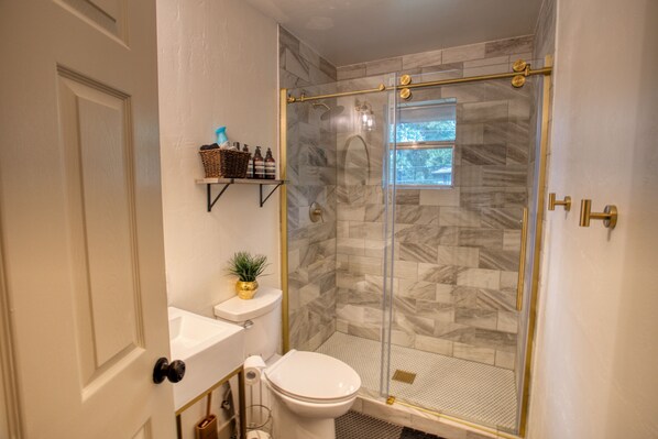 A bathroom with a glass-enclosed shower, a toilet, a sink, and a small shelf with toiletries. - A cozy treehouse cabin in the heart of Pinetop. (Pinetop-Lakeside)