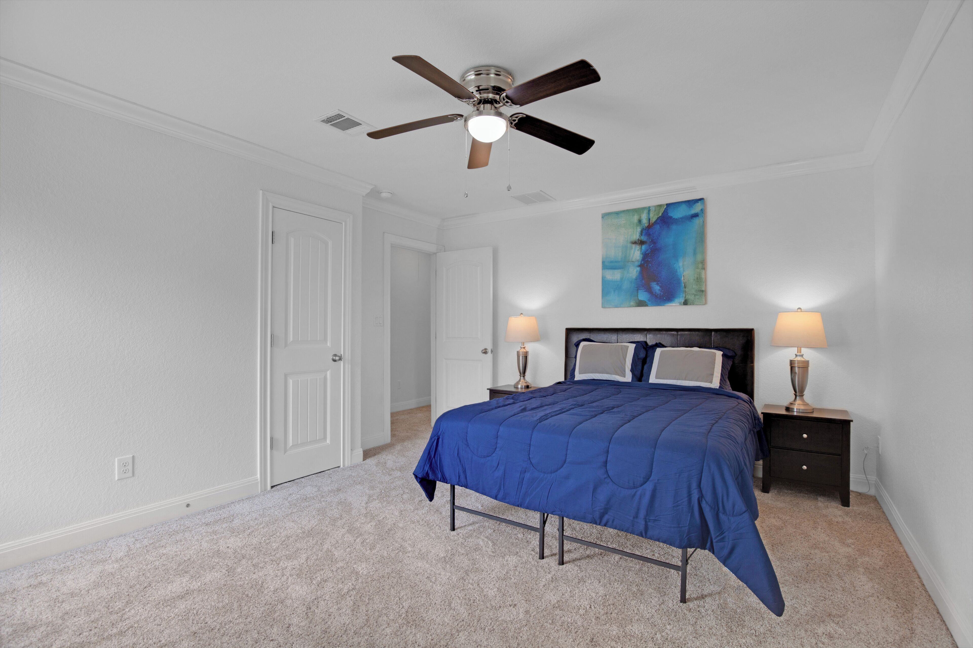 A bedroom with a bed, two bedside tables with lamps, a ceiling fan, a door, and a painting on the wall.