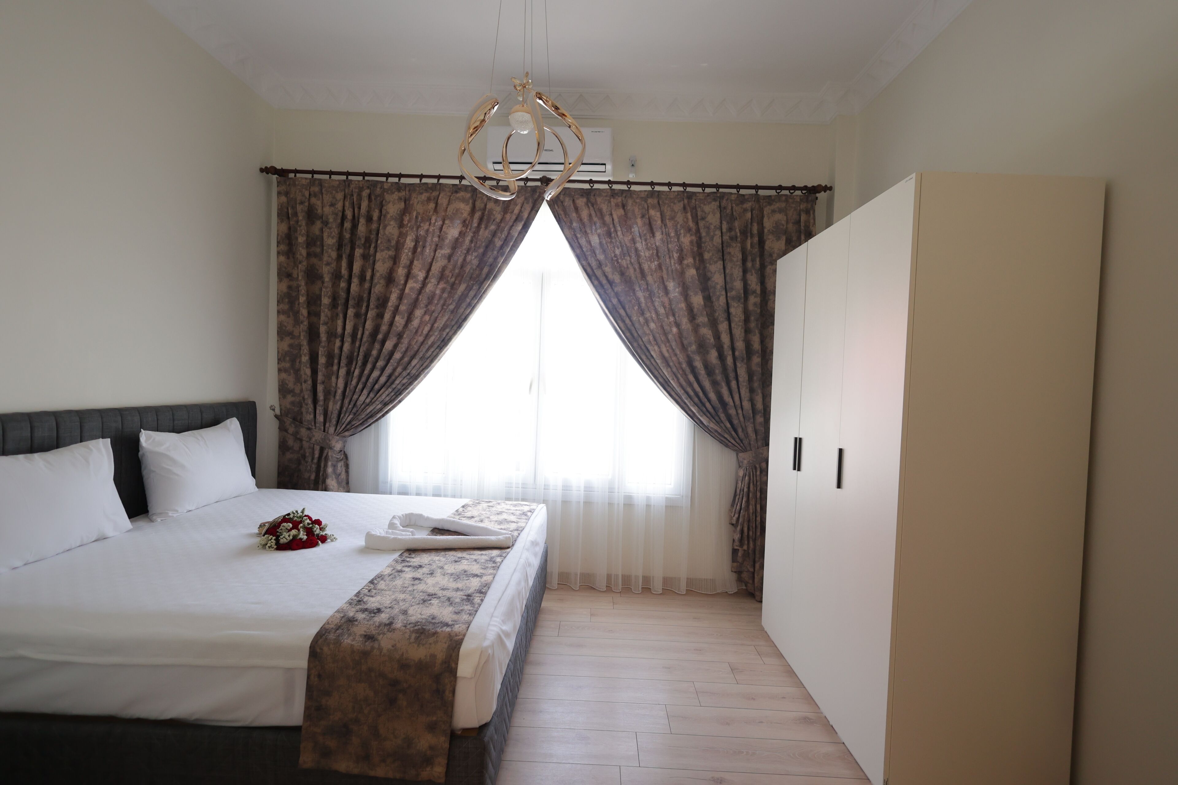 Une chambre d’hôtel avec un grand lit, une table de chevet, un lustre, une fenêtre avec des rideaux et une armoire.
