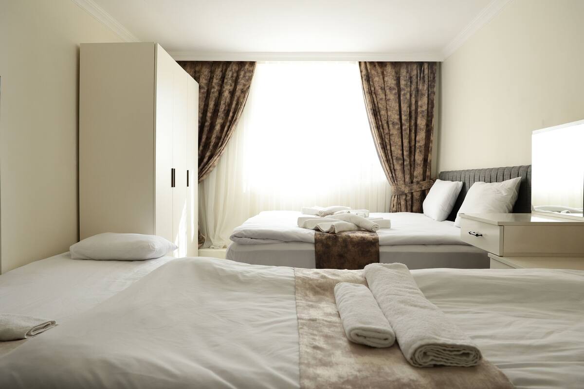 Una habitación de hotel con dos camas, un ventanal con cortinas, una mesita de noche con lámpara y un armario.